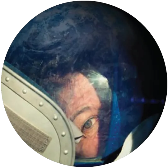 Part of an astronaut's face, including one eye, seen through a porthole window with the blue globe of the Earth reflected in it.